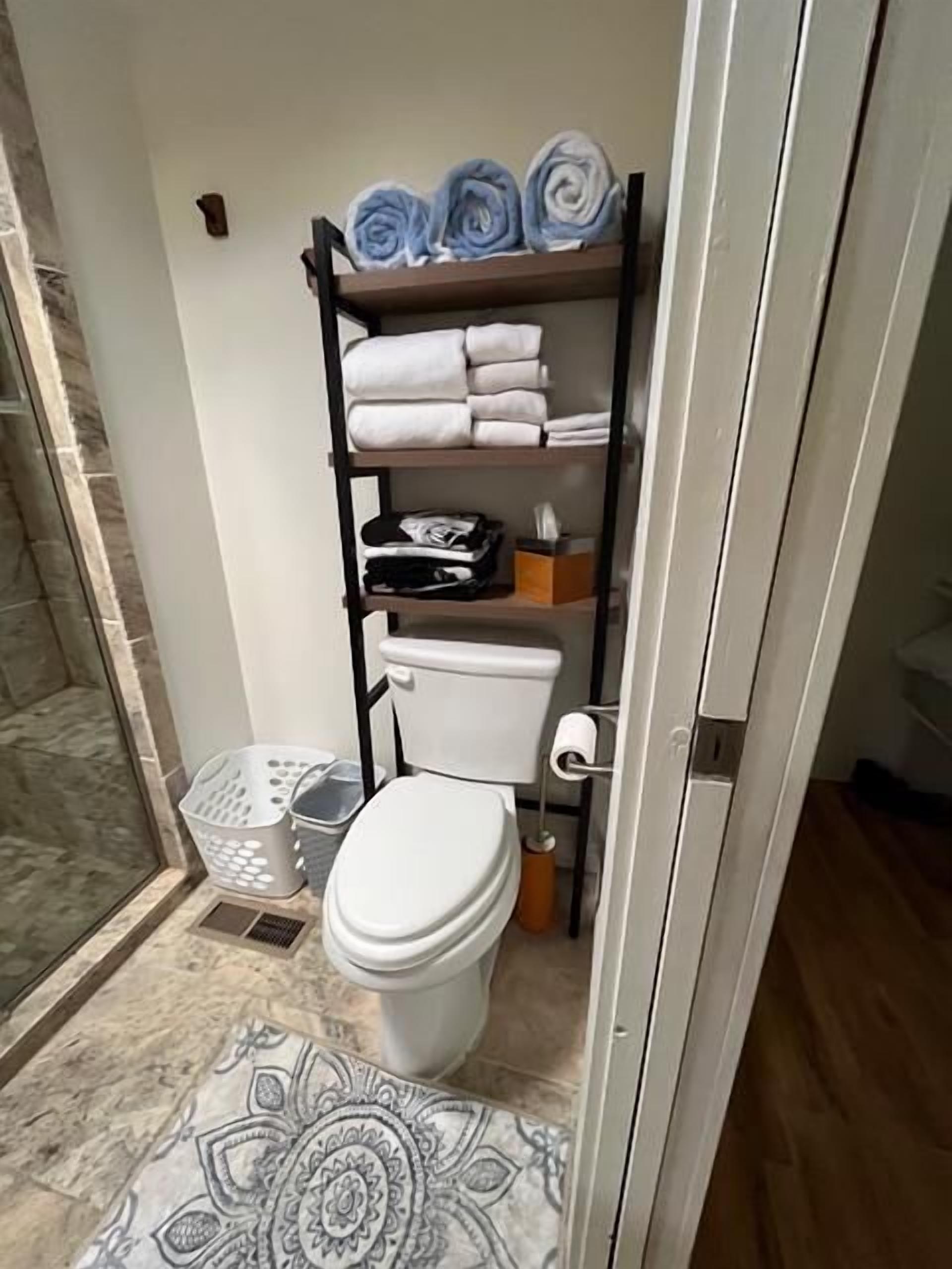 A compact bathroom with a toilet, a shelf with towels, a basket, and a patterned floor.