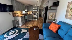 Ein gemütlicher, kompakter Wohnbereich mit Küchenzeile, einem blauen Sofa mit orangefarbenem Kissen und einem gemusterten Teppich.