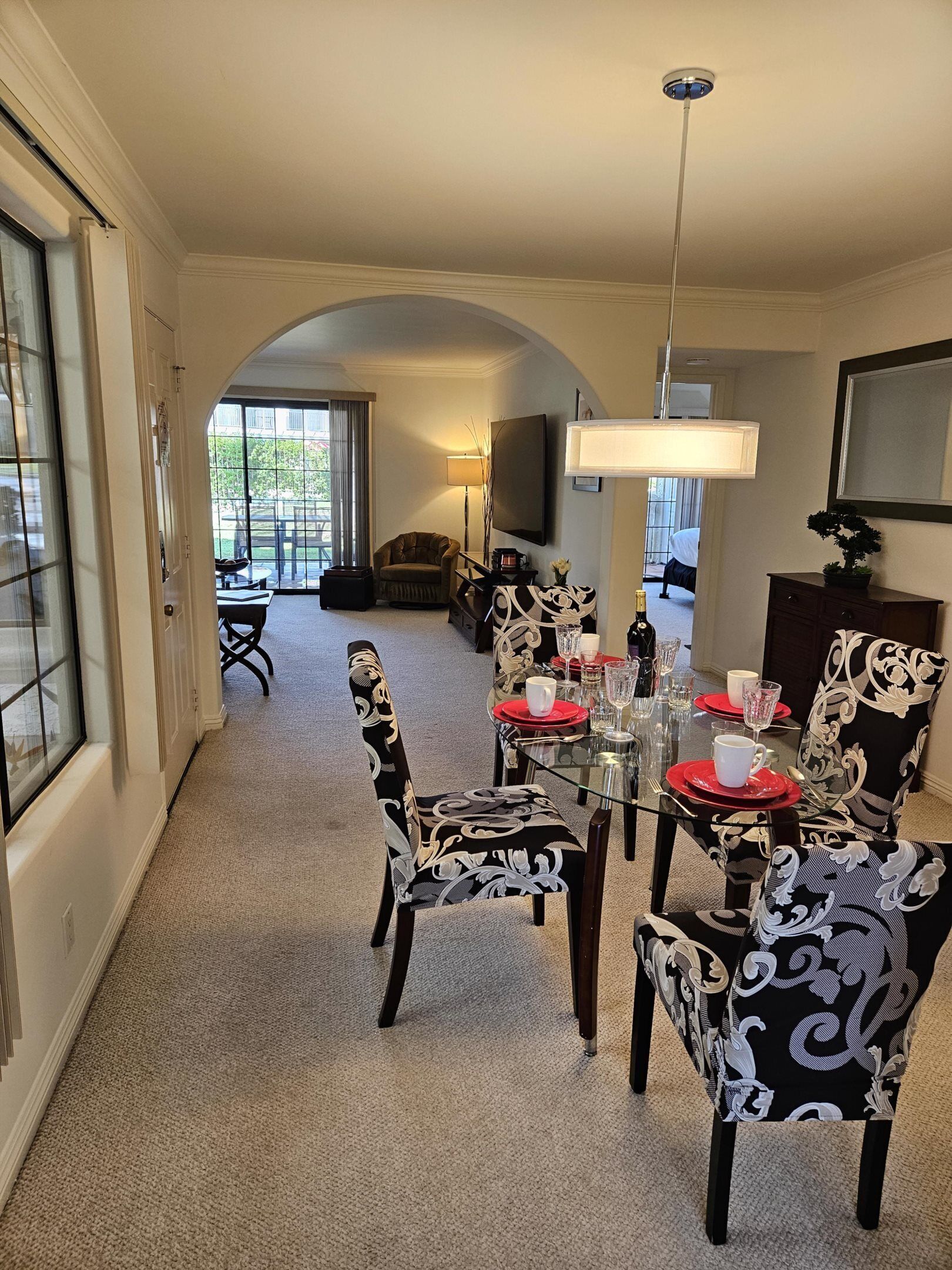 A dining area with a glass table set for four, surrounded by patterned chairs. A sofa and a television are visible in the background.