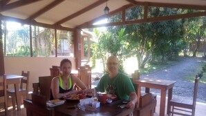 Two people seated at a wooden table with food and drinks, in a covered outdoor area with natural surroundings. - LUMARA LODGE (Guayabo)