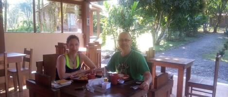 Two people seated at a wooden table with food and drinks, in a covered outdoor area with natural surroundings.