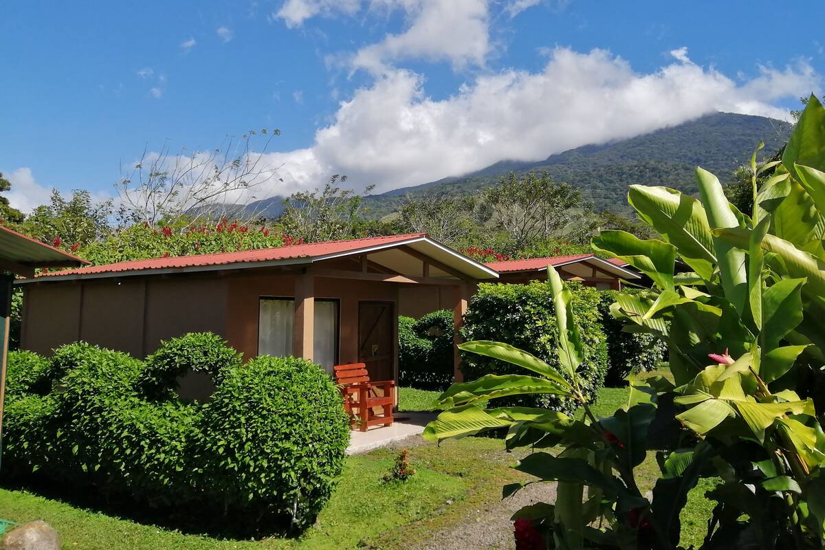 A bungalow with a red roof, surrounded by lush greenery and a mountain backdrop.