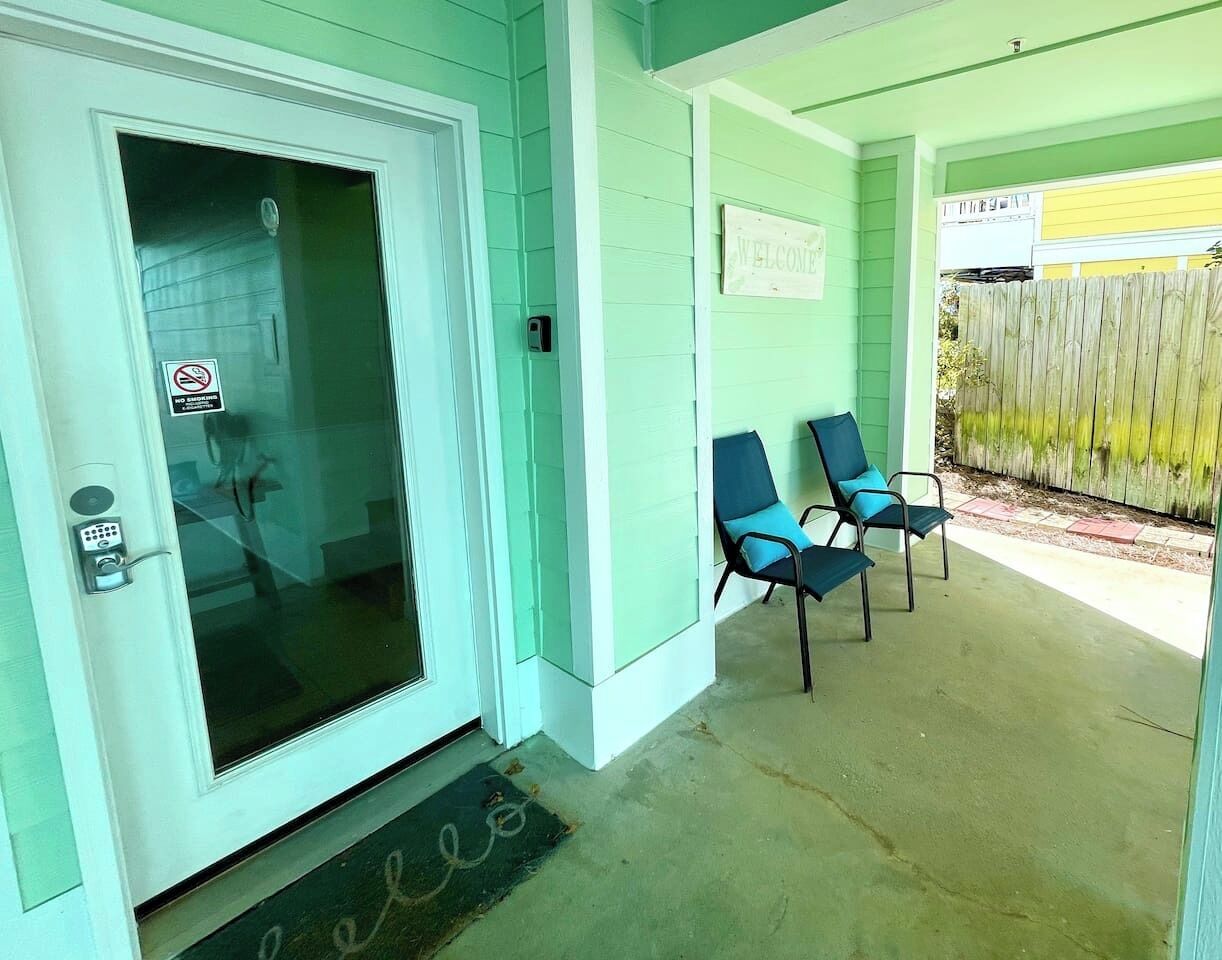 En bygningsindgang i teal farve med en glasdør, to blå stole udenfor, og et skilt med teksten 'Welcome'.