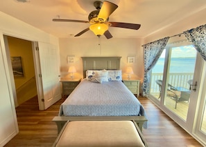 A bedroom with a bed, a ceiling fan, two bedside tables with lamps, a window with curtains, and a view of the water. - Bay Breeze: Gorgeous Waterfront Unit with Kayaks! (Pensacola)