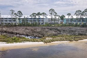Un hôtel situé sur la côte, composé de bâtiments à plusieurs étages, donnant sur la plage et bordé d’une zone marécageuse où poussent de hautes herbes.