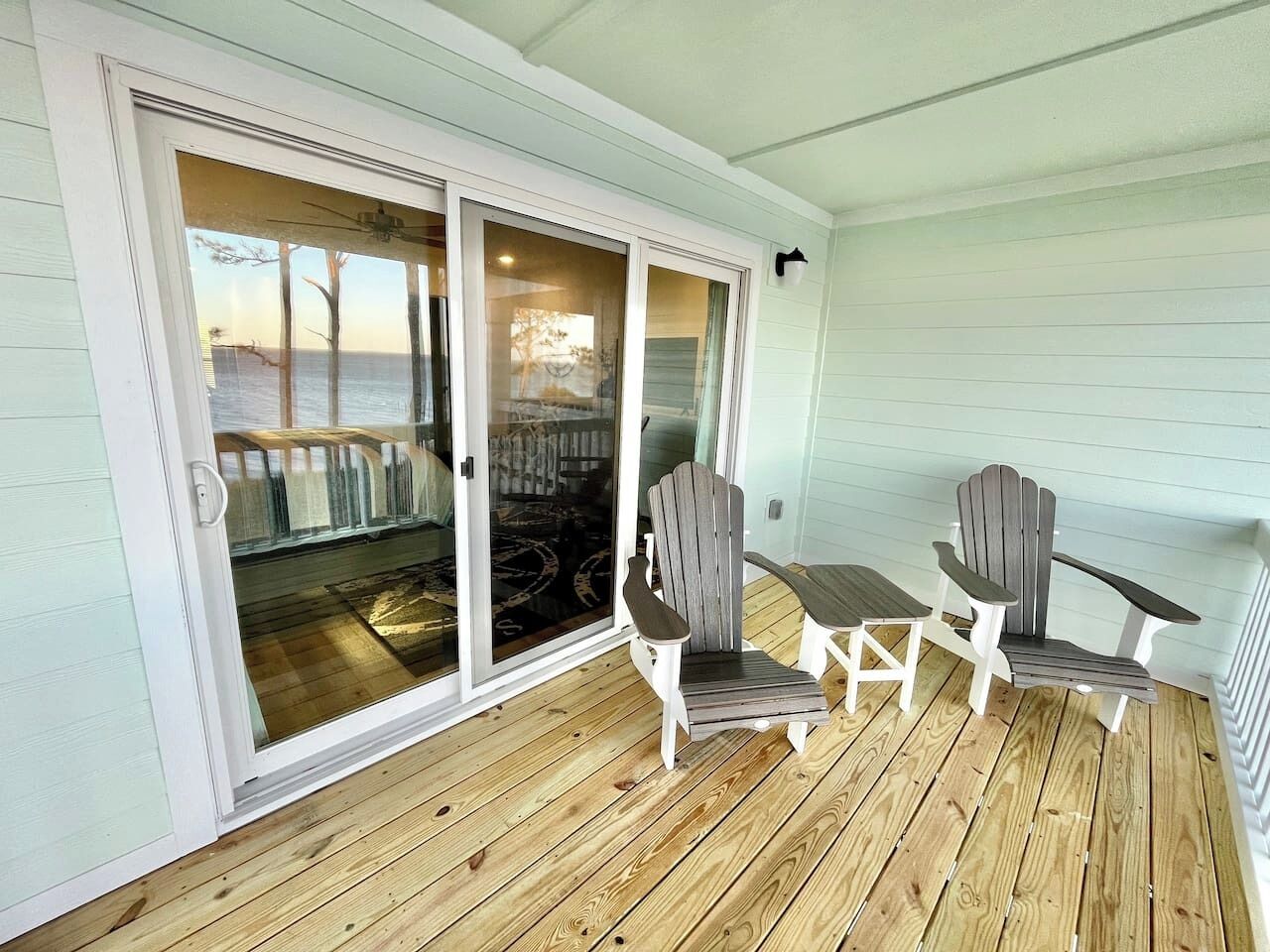 Une terrasse en bois couverte, avec deux fauteuils Adirondack et une petite table, une porte-fenêtre coulissante donnant sur la terrasse, et un ventilateur de plafond.