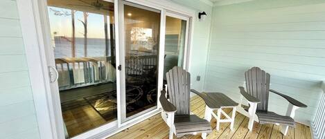 Une terrasse en bois couverte, avec deux fauteuils Adirondack et une petite table, une porte-fenêtre coulissante donnant sur la terrasse, et un ventilateur de plafond.