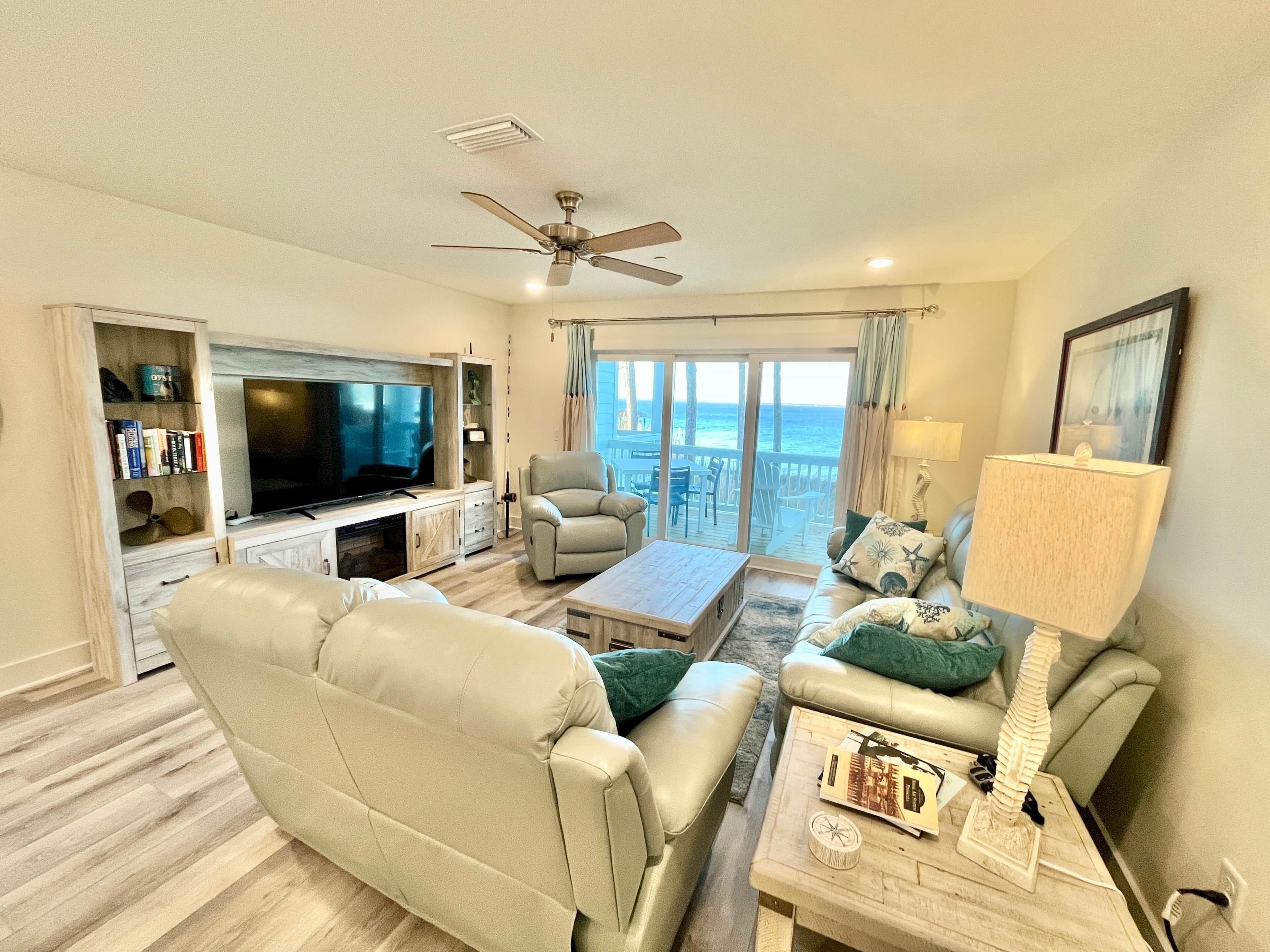 A living room with a television, leather sofa, armchair, and a coffee table. There is a balcony with a view of the ocean.