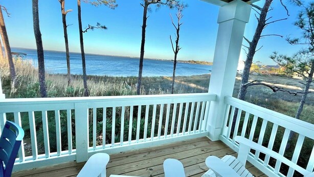 A covered porch with white railings and wooden flooring, overlooking a beach with trees and the ocean.