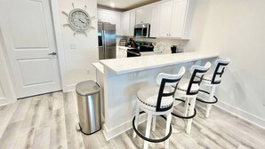 A modern kitchen with white cabinets, a stainless steel refrigerator, a microwave, and a sink. There are three white bar stools with black accents. A trash can is visible to the left of the kitchen.