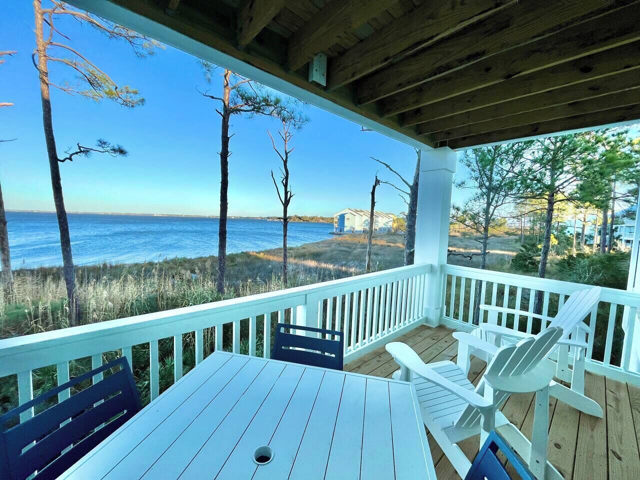 Um deck de madeira com guarda-corpos brancos, uma mesa e duas cadeiras Adirondack com vista para uma praia e oceano.