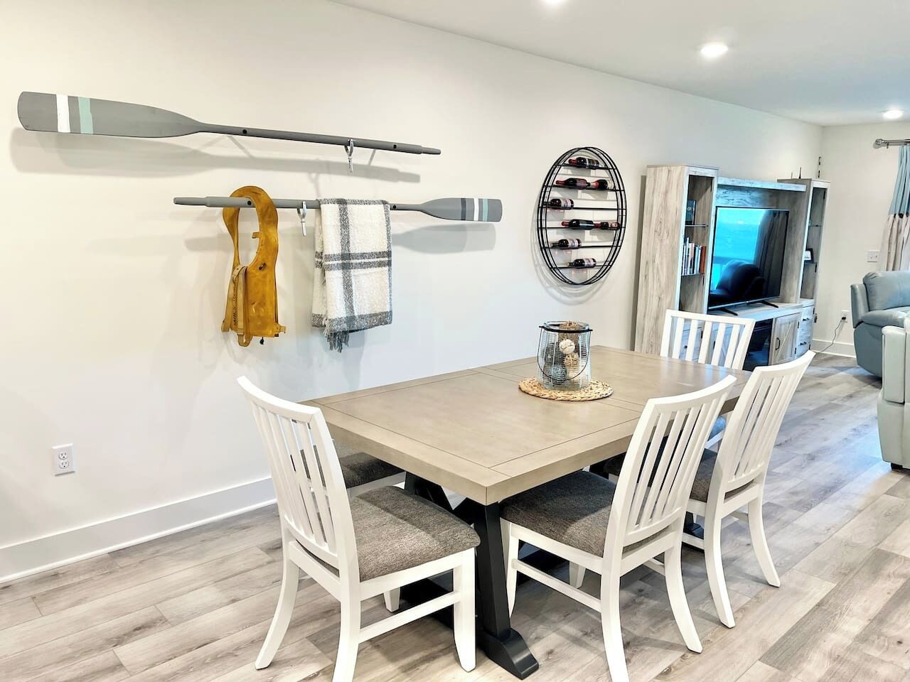 Un comedor con mesa de madera y sillas blancas, una estantería para vinos y un televisor al fondo.