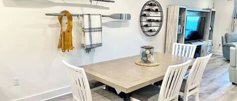 Un comedor con mesa de madera y sillas blancas, una estantería para vinos y un televisor al fondo.