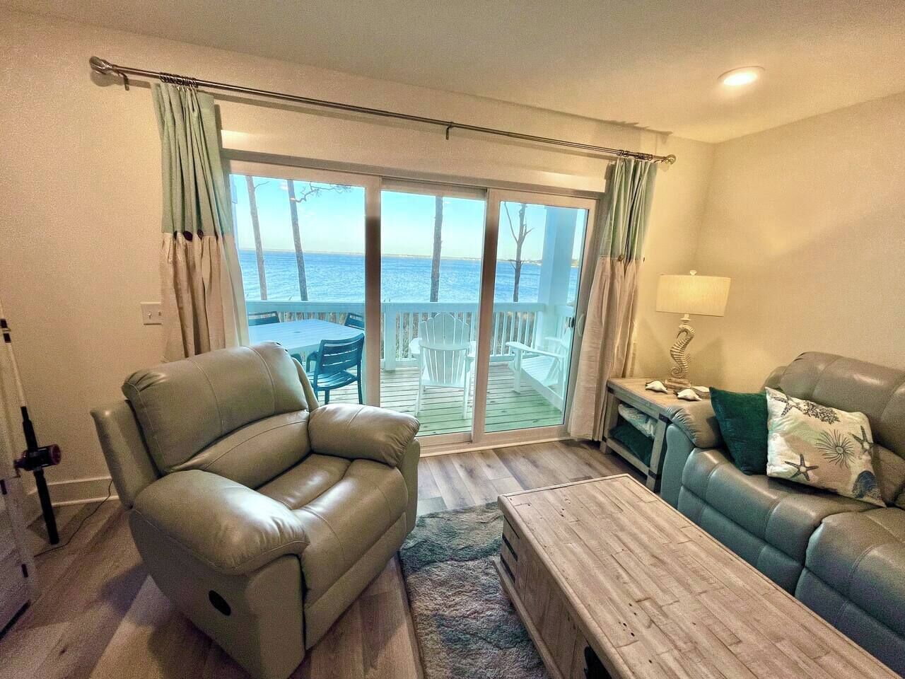 A living room with a beige leather sofa, a blue sofa, and a wooden coffee table. There is a view of the sea through large glass doors.