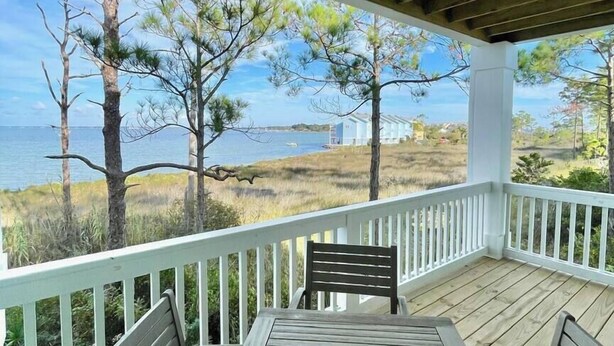 A wooden deck with a table and chairs, overlooking a beach with trees and houses in the distance.