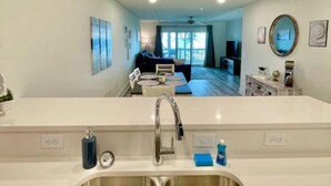 A modern kitchen with a double sink, a dining area with a table and chairs, and a living room with a sofa and a TV.