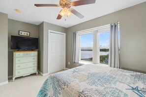 A bedroom with a bed, a dresser, a television, a ceiling fan, a window with a view of a body of water, and a door.