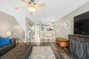 A living room with a ceiling fan, a TV, a sofa, a dining area with chairs, and a kitchen in the background.