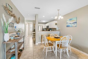 A dining area with a wooden table and white chairs, a kitchen with white cabinets, and a hallway leading to another room.