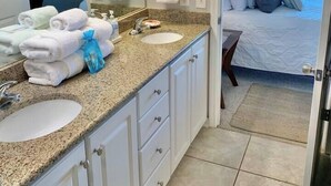 A hotel bathroom with two sinks, a large mirror, and a view of the bedroom through an open door. - Serenita Ora: Quiet Waterfront Unit with 2 Kayaks! (Pensacola)