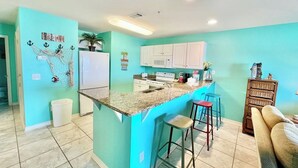 A kitchen with a turquoise accent wall, white cabinets, a granite countertop, and a small island with bar stools. - Tiki Hut: Waterfront, Amazing Views, & 2 Kayaks! (Pensacola)