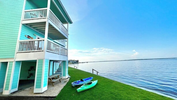 A two-story building with a balcony, a grassy area, and two kayaks. - Tiki Hut: Waterfront, Amazing Views, & 2 Kayaks! (Pensacola)