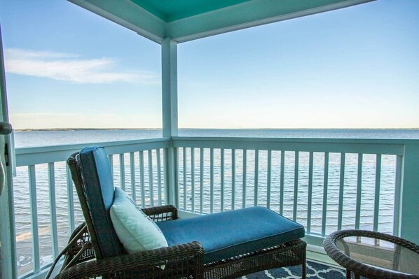 A covered balcony with a wicker chair and a small table, overlooking a calm sea.