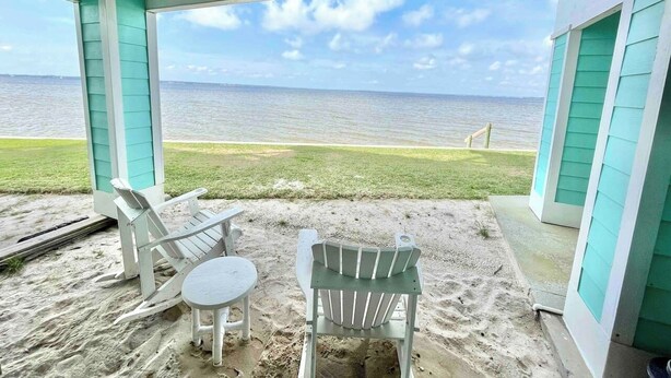 A covered outdoor seating area with two Adirondack chairs and a small table, overlooking a beach with a view of the ocean. - Bay Paradise: Paddle Boards, Kayaks, & Waterfront! (Pensacola)