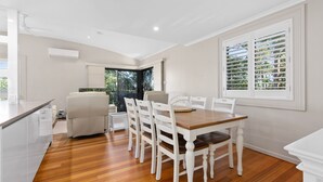 A dining area with a wooden table and white chairs, a white sofa, and a large window with blinds. - 21 Cooloola Drive - Rainbow Beach (Rainbow Beach)