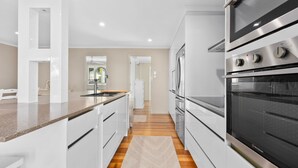 A modern kitchen with white cabinetry, a granite countertop, and stainless steel appliances including a double oven and a refrigerator. - 21 Cooloola Drive - Rainbow Beach (Rainbow Beach)