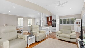 A modern living room with a sofa, armchairs, and a coffee table. There is a kitchen area with appliances and a dining table. - 21 Cooloola Drive - Rainbow Beach (Rainbow Beach)