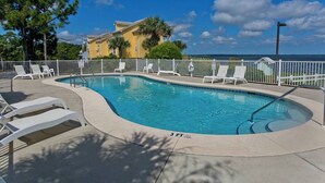 A swimming pool with lounge chairs and a fence, surrounded by trees and a building in the background.