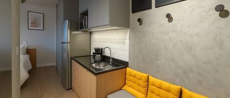 A compact kitchen with a built-in microwave, a coffee maker, and a sink. There is a yellow cushioned bench against the wall and a framed picture of the moon.