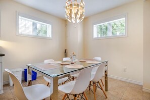 Un coin repas avec une table en verre et des chaises blanches, un lustre moderne et des fenêtres équipées de stores.