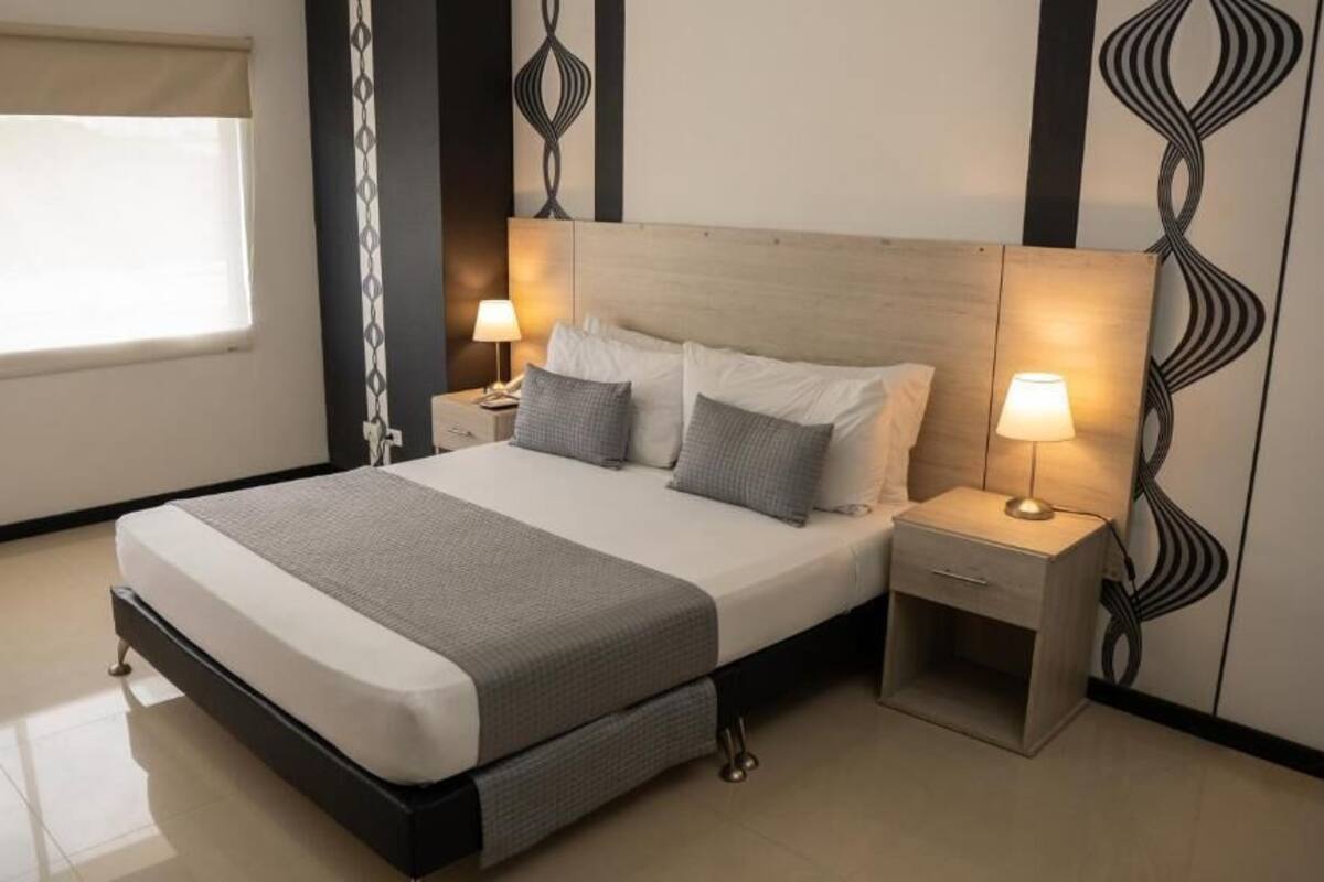Una habitación de hotel moderna con una cama grande, dos mesitas de noche con lámparas, un ventanal con persianas y un diseño de pared estampado.