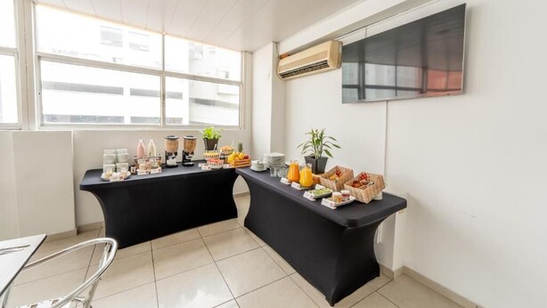 A breakfast buffet setup with various food items and beverages on black tables, a potted plant, and a mounted TV on the wall. - Basic Hotel Chipichape By Hoteles MS (Cali)