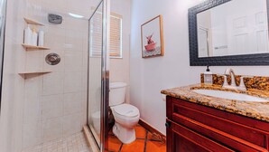 Un baño con ducha envuelta en vidrio, inodoro, lavamanos con encimera de granito y un espejo grande con marco negro.