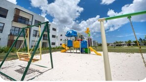 A playground with swings and slides, set against a backdrop of modern buildings and a clear sky. - Punta Cana Beach Retreat-Costa Bavaro (Punta Cana)