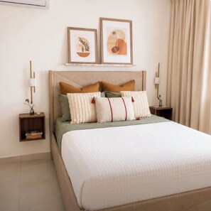 A neatly made bed with a quilted white cover, flanked by two bedside tables with lamps, a small shelf with books, and two framed artworks on the wall. - Punta Cana Beach Retreat-Costa Bavaro (Punta Cana)