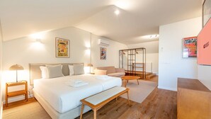 Una habitación de hotel moderna con una cama grande, un banco de madera, un sofá y una mesita pequeña. Hay una escalera con barandilla de vidrio y un cartel en la pared.