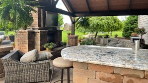 Eine Terrasse im Freien mit Kamin aus Stein, Korbmöbeln und einer Arbeitsplatte aus Granit.
