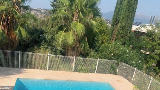 A rectangular swimming pool with a tiled deck, surrounded by a metal fence and lush greenery, including palm trees and a tall coniferous tree. - Bel air flat (Villeneuve-Loubet)