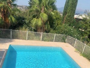 En rektangulær swimmingpool med fliseterrasse, omgivet af et metalhegn og frodig beplantning, herunder palmer og et højt stedsegrønt træ.