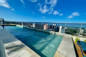 Une piscine sur le toit, avec une eau d’un bleu limpide, entourée d’une barrière de verre, offrant une vue imprenable sur le paysage urbain et la mer.