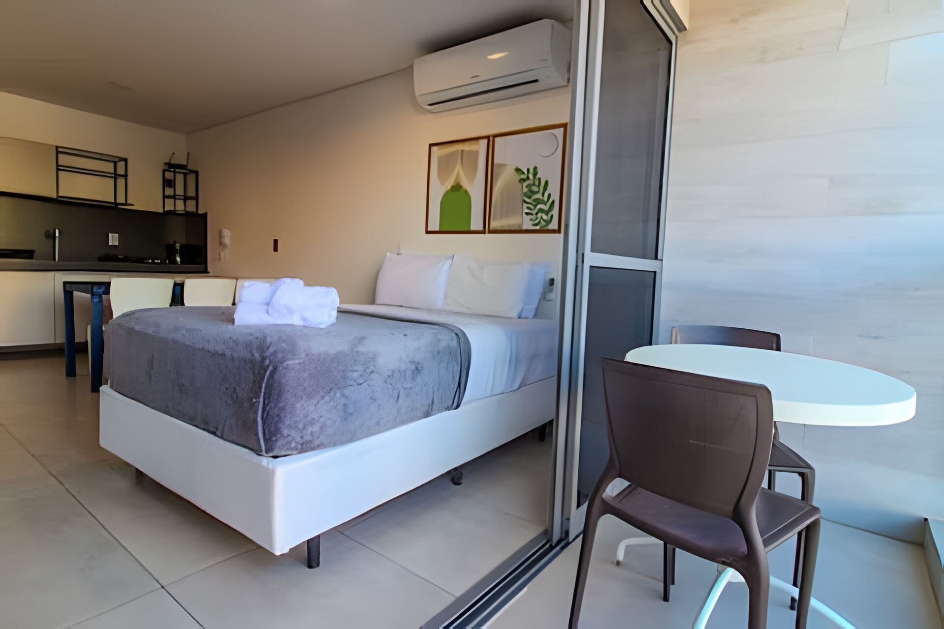 A modern hotel room with a bed, a small dining table, and a kitchenette in the background.