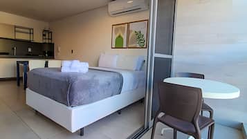 A modern hotel room with a bed, a small dining table, and a kitchenette in the background.