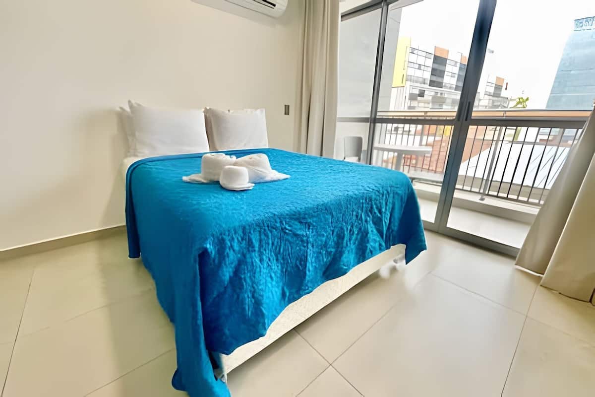 Une chambre d’hôtel avec un grand lit, des oreillers blancs, une couvre-lit bleue et une vue sur les immeubles depuis un balcon.