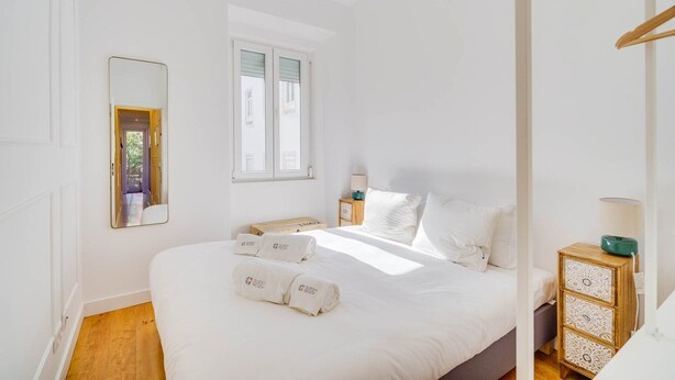 A bright, white room with a large bed, a mirror, a window, and a bedside table with a lamp. - GuestReady - Relaxing place in Lisbon (Lisbon)