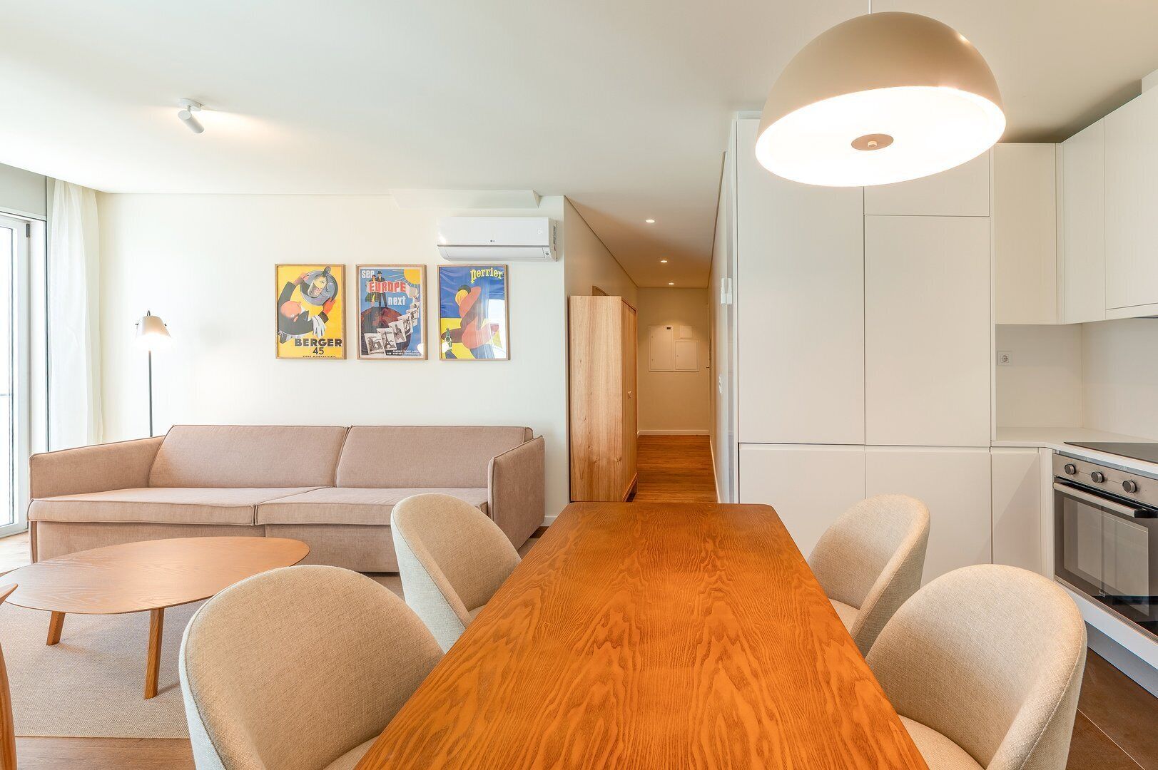 Una sala de estar moderna con una mesa de comedor de madera, un sofá color beige y una cocina con gabinetes blancos y una estufa.