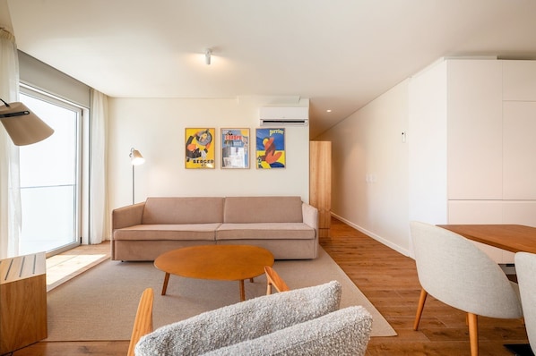 A modern living room with a sofa, a round coffee table, and a dining table with chairs. There are three framed artworks on the wall and an air conditioning unit above the sofa.